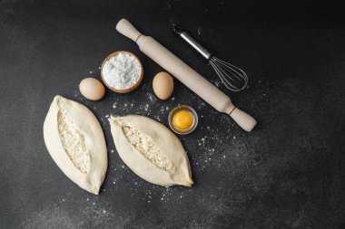 İki çiğ khachapuri ekmeği, içinde fırın malzemeleri olan siyah un kaplı bir masada.