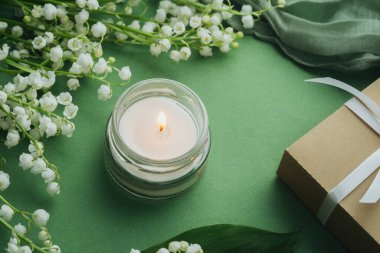 Yeşil arka planda beyaz zambaklı cam mum yanıyor.