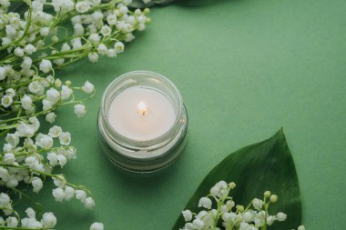 Yeşil arka planda beyaz zambaklı cam mum yanıyor.