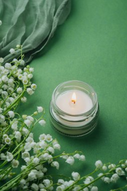 Yeşil arka planda beyaz zambaklı cam mum yanıyor.