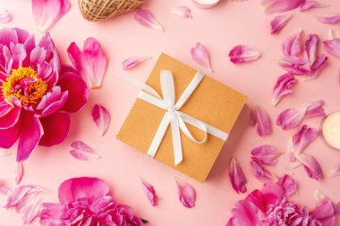 Beyaz kurdeleli hediye kutusu şakayıklar, taç yaprakları, mumlar ve pembe arka planda sicimle çevrili.