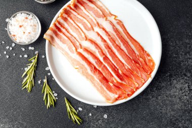 Beyaz tabakta çiğ domuz pastırması, biber, tuz ve biberiye.