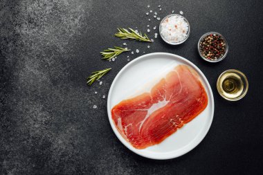 Beyaz tabakta dilimlenmiş jambon ve siyah arka planda baharatlar.