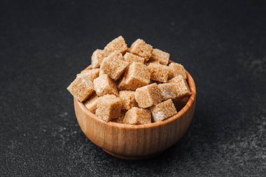 Koyu bir yüzeye yerleştirilmiş küçük kahverengi şeker küpleriyle dolu ahşap bir kase..