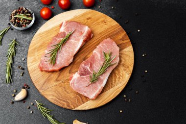 Çiğ domuz pirzolası, taze biberiye ve vişneli domateslerle ahşap kesim tahtasına konmuş..