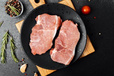 İki çiğ domuz pirzolası siyah bir tabağa konur. Etrafı otlar ve baharatlarla çevrilidir..