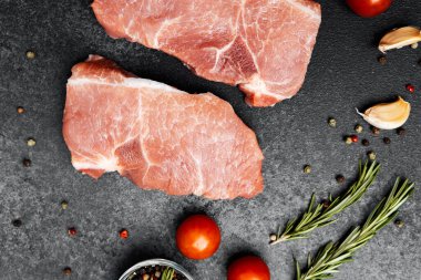 İki pişmemiş domuz pirzolası domates, sarımsak ve karanlık bir yüzeye yerleştirilmiş otlar..