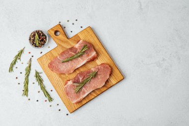 Yanında biberiye ve baharat olan bir kesim tahtasının üzerinde iki çiğ domuz pirzolası..