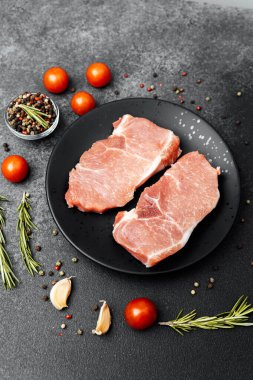 Baharat, biberiye ve domateslerle çevrili bir tabakta iki çiğ domuz pirzolası..