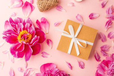 Beyaz kurdeleli hediye kutusu şakayıklar, taç yaprakları, mumlar ve pembe arka planda sicimle çevrili.