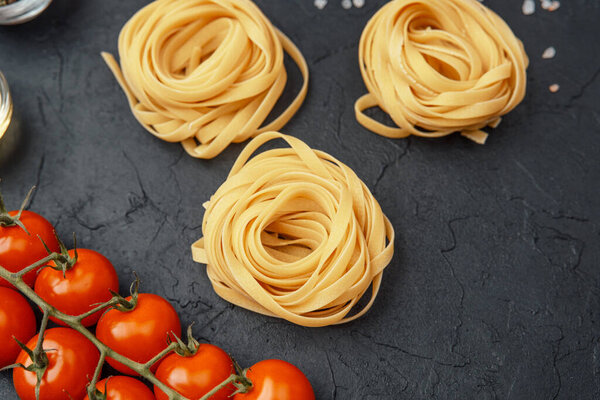 Tagliatelle pasta nests with tomatoes, oil, pepper and salt on black textured background