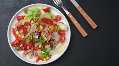 Taze ton balığı salatası, karışık sebzeler ve zeytinlerle servis edilir. Beyaz bir tabakta, yakınlardaki karanlık bir yüzeye yerleştirilmiş çatal ve bıçakla..