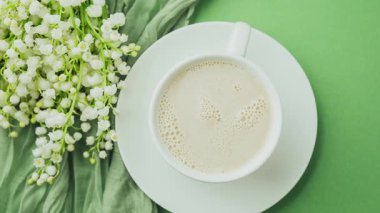 Basit bir beyaz bardak ılık köpüklü içecek narin beyaz çiçekler ve yumuşak yeşil kumaşın yanında yeşil bir arka planda yatıyor..