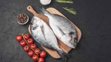 Taze çiğ balık, olgun domatesler, taze otlar ve koyu bir zemin üzerinde sağlıklı deniz ürünleri hazırlamak için baharatlarla ahşap bir kesme tahtasının üzerinde yatar..