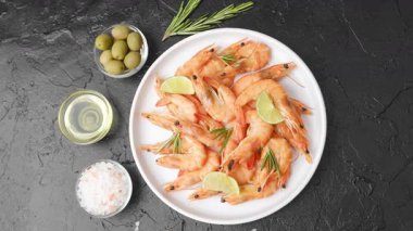 Beyaz tabakta limonlu ve biberiyeli nefis pişmiş karides. Yeşil zeytin, yağ ve tuz koyu dokulu bir zemin üzerindedir..