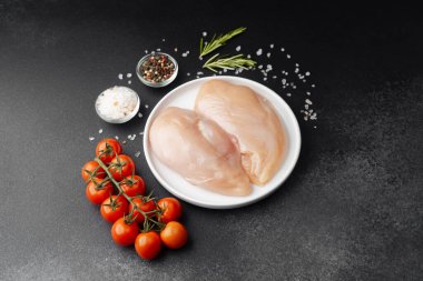 Beyaz tabakta çiğ tavuk filetosu, siyah masada tuz, domates ve otlar.