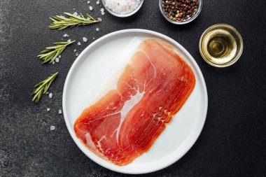 Beyaz tabakta dilimlenmiş jambon ve siyah arka planda baharatlar.