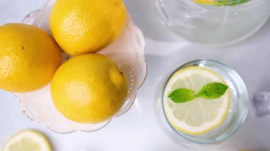Taze sarı limonlar ve taze yeşil nane yapraklı ferahlatıcı bir bardak limon suyu temiz beyaz bir yüzey üzerinde güzelce sergilenir..