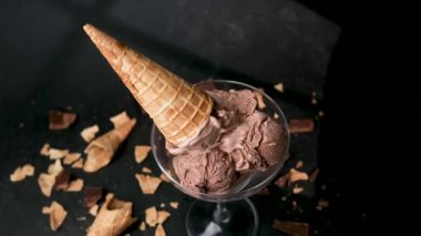 Zengin çikolatalı dondurma kepçeleri waffle külahlı bir bardakta servis edilir ve koyu bir arka planda dağınık parçalar..