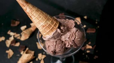 Çikolatalı dondurma kepçeleri bir cam kasenin içinde, tatlıya koni konulmuş, dağınık kırıntıları olan koyu bir arka planda..