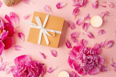 Beyaz kurdeleli hediye kutusu şakayıklar, taç yaprakları, mumlar ve pembe arka planda sicimle çevrili.