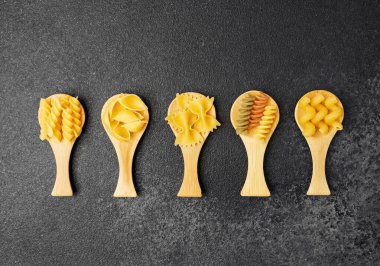 Koyu renkli bir yüzeyde ahşap kepçelerle sıralanmış çeşitli makarna türleri..