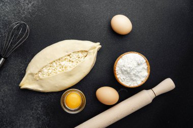 Süzme peynirli çiğ khachapuri ve siyah arka plandaki malzemeler.
