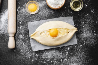 Çiğ khachapuri, yumurta sarısı ve parşömen üzerinde peynir ve siyah arka planda un.