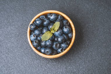 Koyu bir yüzey üzerinde nane yapraklarıyla ahşap bir kasede dizilmiş taze yaban mersini..