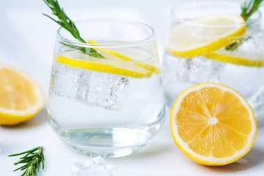 Temiz bir bardak limon suyu biberiye filizi ve taze limonun yanında buz. Yarısı gün ışığında arkaplanda.