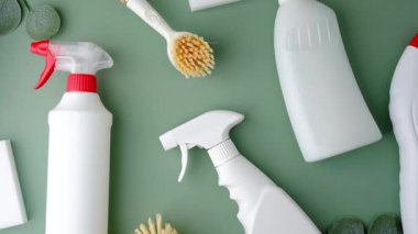 Beyaz sprey şişeleri ve temizlik fırçası yeşillik detaylarıyla sade yeşil bir arka planda düzenlenmiş.