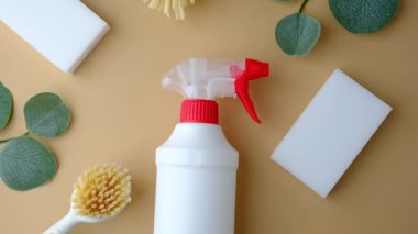 Yeşil yaprakların yanında kırmızı kapaklı beyaz sprey şişesi ve açık kahverengi yüzeyde temizlik aletleri..