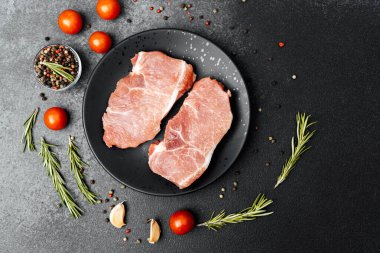 Bitkiler, domatesler ve baharatlarla çevrili bir tabakta iki çiğ domuz pirzolası..