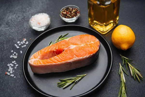 Yakındaki taze otlar, limonlar ve baharatlarla siyah bir tabağa konmuş somon filetosu..