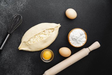 Süzme peynirli çiğ khachapuri ve siyah arka plandaki malzemeler.
