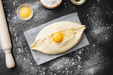Çiğ khachapuri, yumurta sarısı ve parşömen üzerinde peynir ve siyah arka planda un.