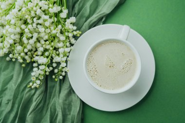 Sütlü bir fincan kahve ve yeşil tekstil arka planında vadi çiçekleri.