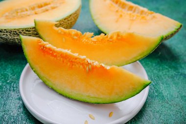 Bir tabakta taze kavun dilimleri, parlak turuncu et ve tohumları gösteriyor. Yeşil desenli yüzey.