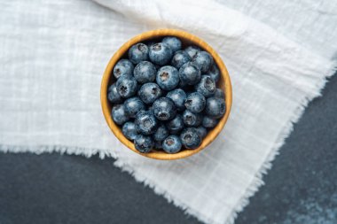 Olgun yaban mersiniyle dolu bir kase dokulu beyaz bir bez yüzeyde duruyor..