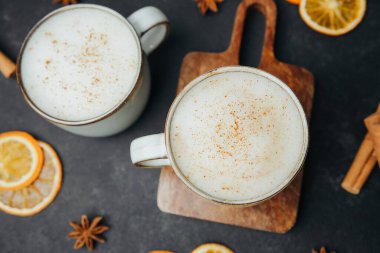 Üzerine tarçın serpiştirilmiş köpüklü içecekler serpiştirilmiş iki sıcak kupa turuncu dilimler ve tarçın çubuklarıyla karanlık bir yüzeye oturur..