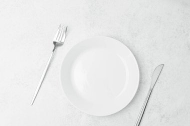 Beyaz bir tabak, çatal ve bıçağın yanında, açık bir yüzeyde duruyor..