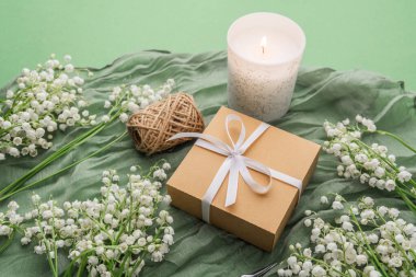 Yeşil kumaş arka planında hediye kutusu olan sicim, mum ve çiçekler.