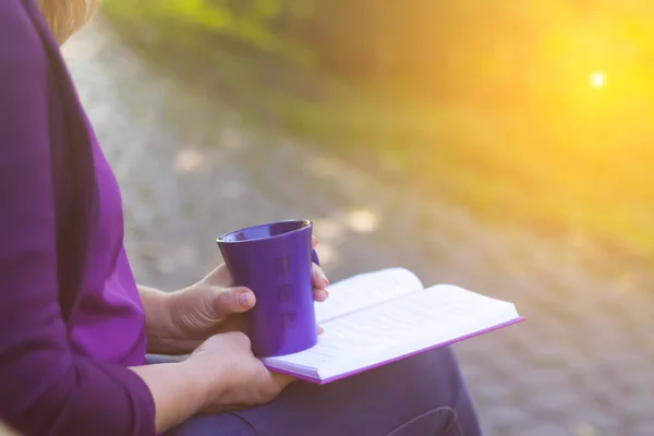 Defocus of woman hand holding and reading a book with holding purple mug cup of hot coffee. A woman sits near a tree in an autumn forest and holds a book and a cup in her hands. Out of focus.