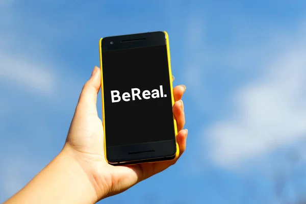 Mavi gökyüzü arka planında sarı plastik çantalı akıllı telefon. Bereal uygulaması. BeReal yeni bir sosyal medya uygulaması ve fotoğraf paylaşım platformu. Volodymyr, Ukrayna, 23 Ekim 2022.