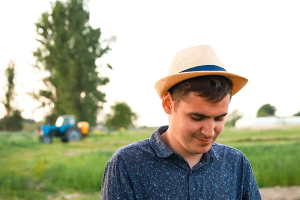 Defocus farmer in hat working on field with greenhouse outdoor. Portrait of young Caucasian handsome happy man farmer standing in field and smiling. Tractor on background. Out of focus.