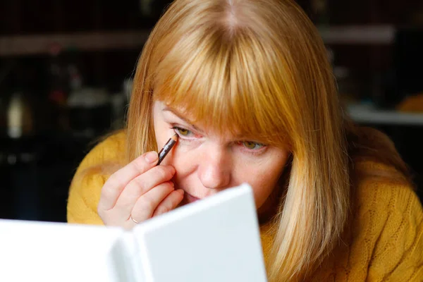 Blonde woman making up eyelashes herself with a hand mirror indoor. Eyeliner makeup. Attractive blonde woman putting makeup. Woman looks in mirror using cosmetic pencil. Out of focus.