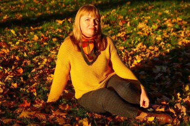 Defocus portrait of young Caucasian blonde woman in autumn park. Bright stylish woman in yellow pullover and shawl of neck seating in park. Happy fall concept. Sunny weather, sunset. Out of focus.