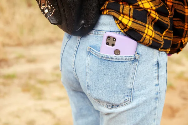 Woman puts mobile phone in jeans pocket on yellow nature background. Blue denim. Smart phone in the purple case. Out of focus.