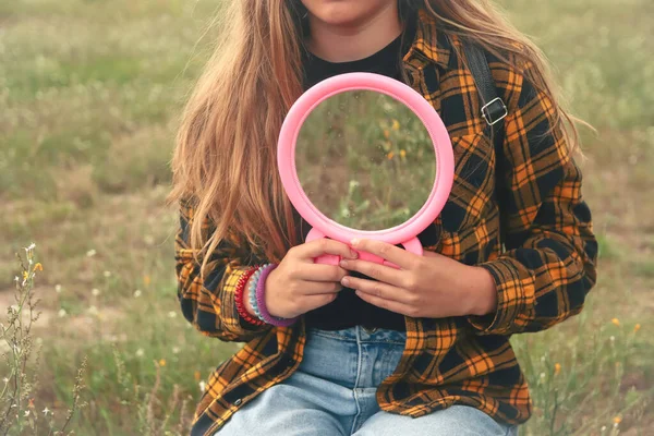 Young girl holding mirror on the nature green background. Beauty nature concept. Grass reflection in round pink mirror on summer field. Floral spring background. Eco concept. Out of focus.