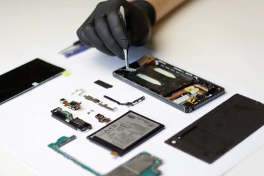 Parçalanmış bir akıllı telefonun profesyonel onarımı, kırık parçalar üzerinde çalışan cımbız ve eldivenli usta. Parçalanmış akıllı telefon parçaları yerleştirilmiş..